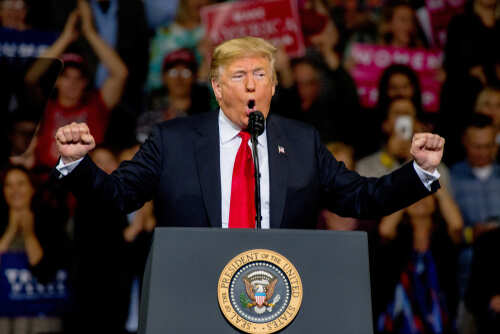 shutterstock_1204270501.jpg A man in a suit passionately speaking at a podium with a presidential seal, surrounded by a cheering crowd