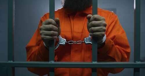 shutterstock_2345749519.jpg A prisoner in an orange jumpsuit holding jail bars with handcuffs